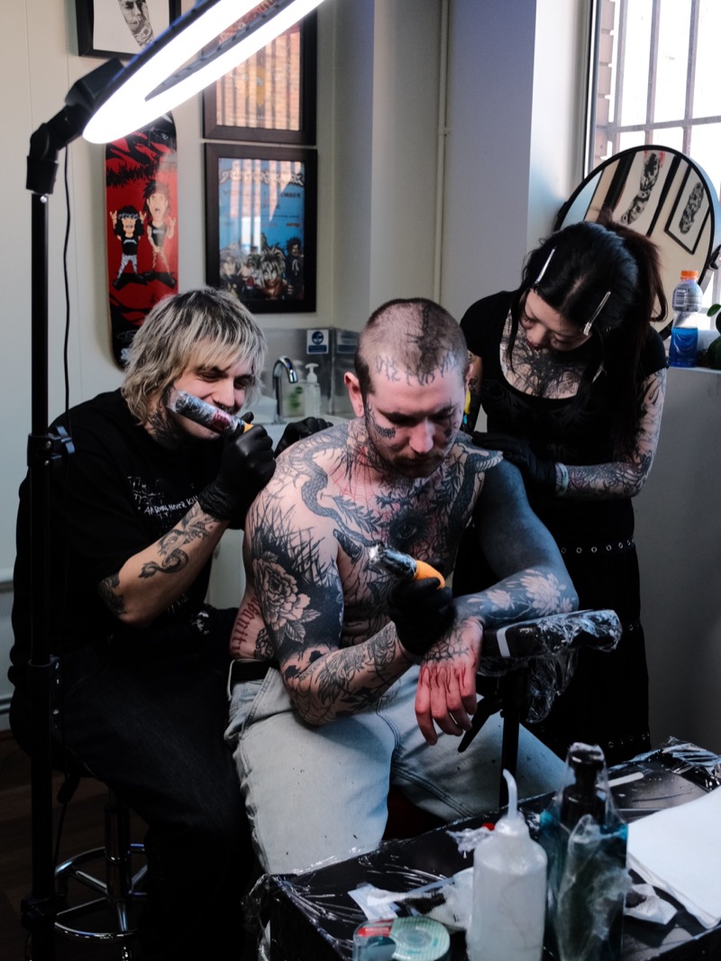 Two artists tattooing a seated client at CAN TATTOO, with framed artwork and ring lights behind them