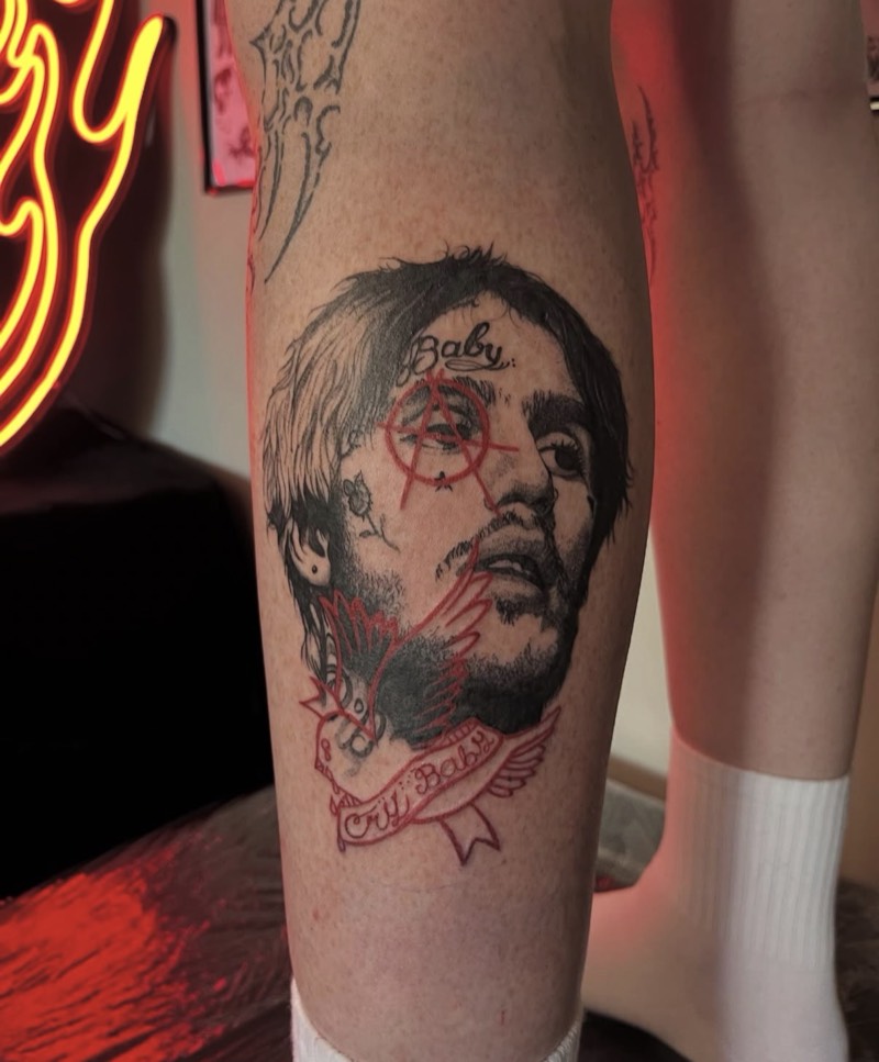 Completed tattoo on the calf: a black and grey portrait of a man with face tattoos, a red anarchy symbol over one eye, and a red bird and banner reading "Cry Baby"
