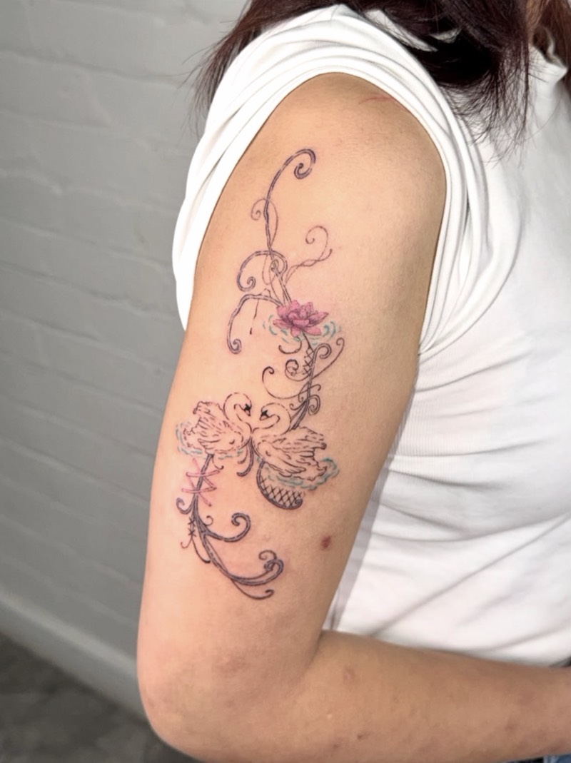 Completed tattoo on the upper arm: two swans on rippling water with a pink lotus and fine curling scrollwork