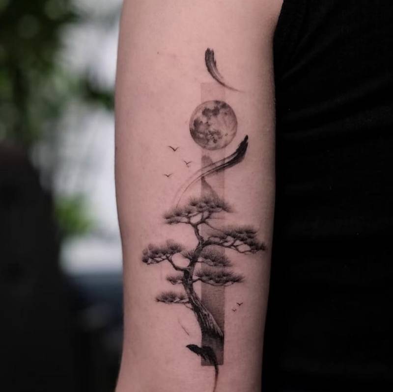 Completed tattoo on the forearm: a pine tree beneath a shaded moon, with small birds and sweeping brush strokes in black and grey