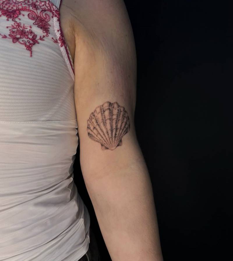 Completed tattoo on the upper arm: a scallop shell with radiating ridges in black stippled shading
