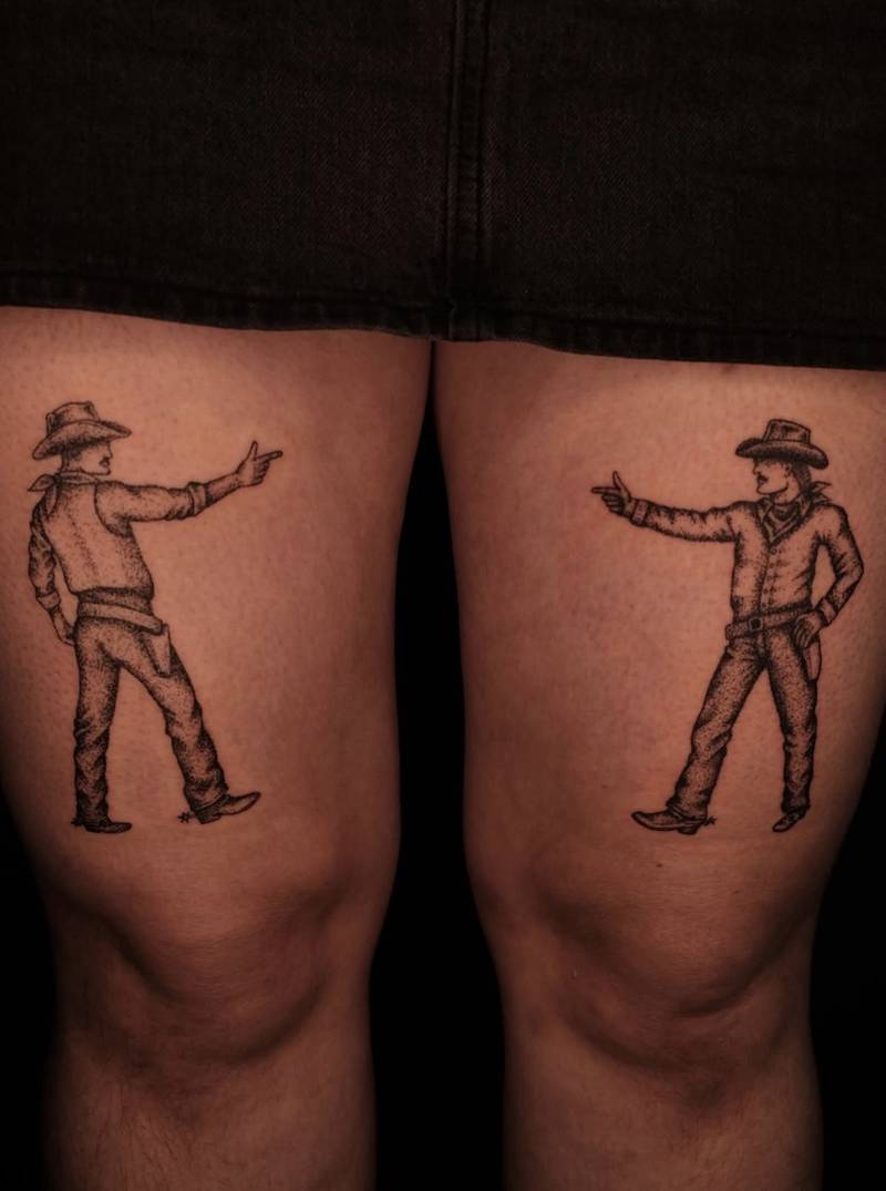 Completed tattoos on the thighs: two cowboys in hats and boots facing one another and pointing with their hands, in black stippled shading
