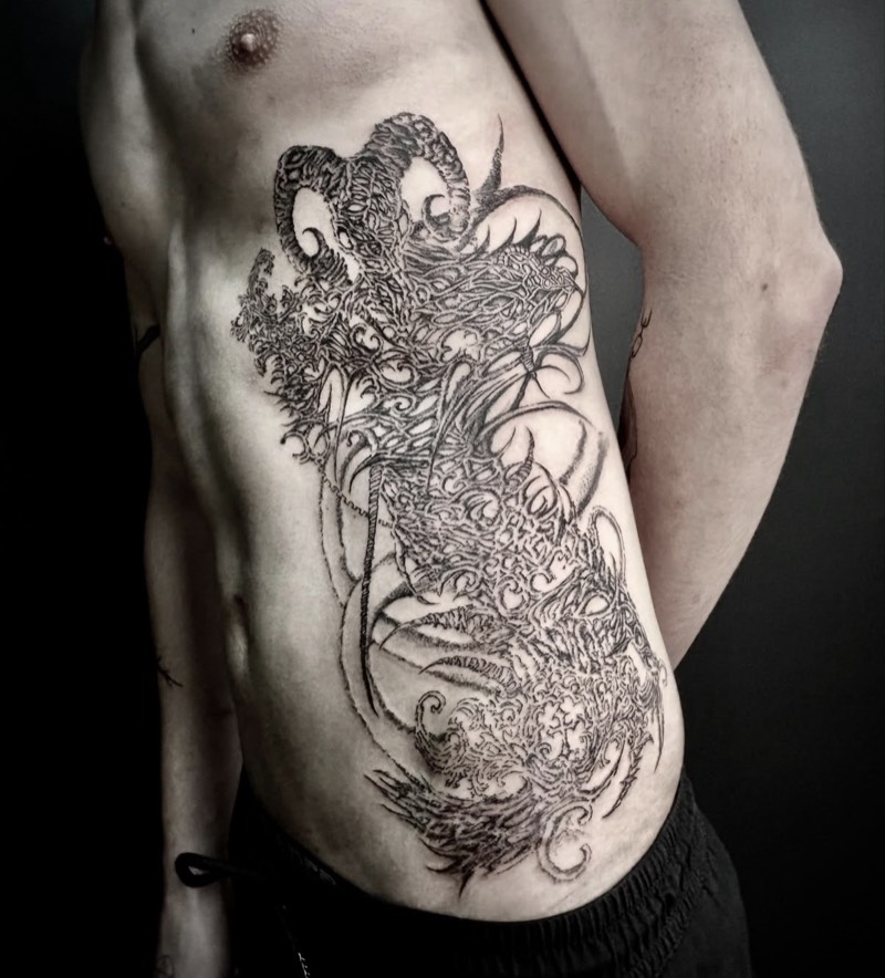 Completed tattoo on the ribs: a large horned ornamental design of dense black linework and curling tendrils