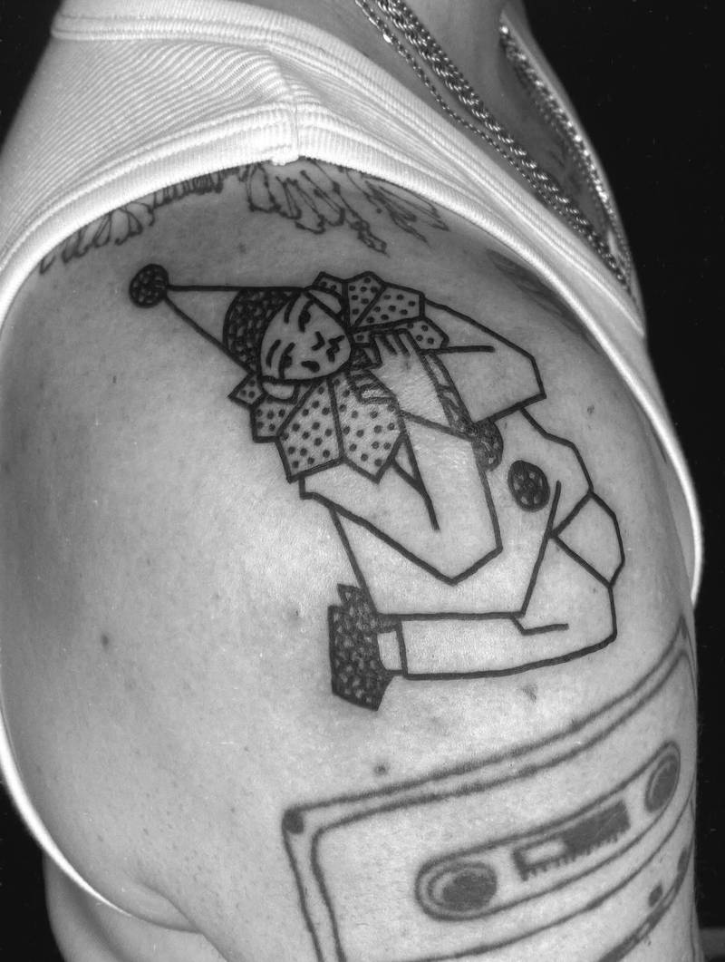 Completed tattoo on the upper arm: a seated clown in a pointed hat and patterned ruff, playing a flute, in black linework