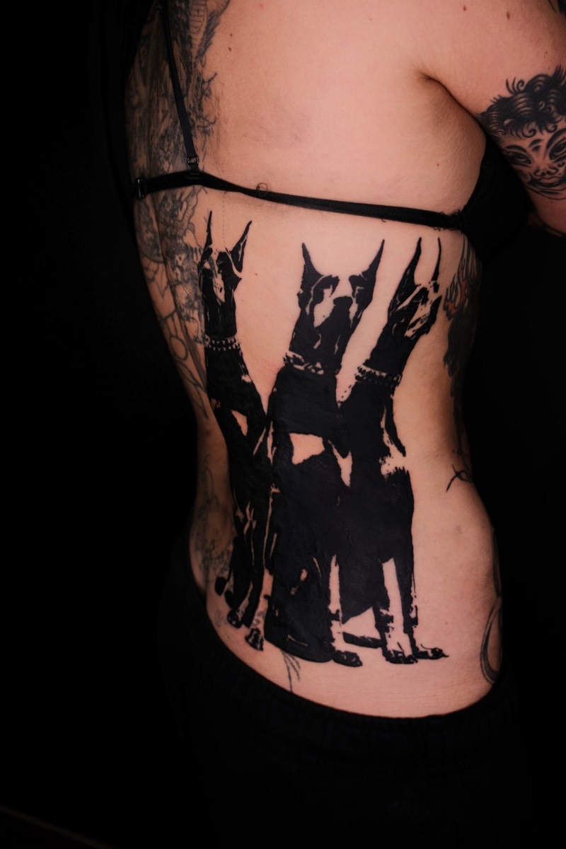 Completed tattoo on the ribs: three seated dobermans in solid black
