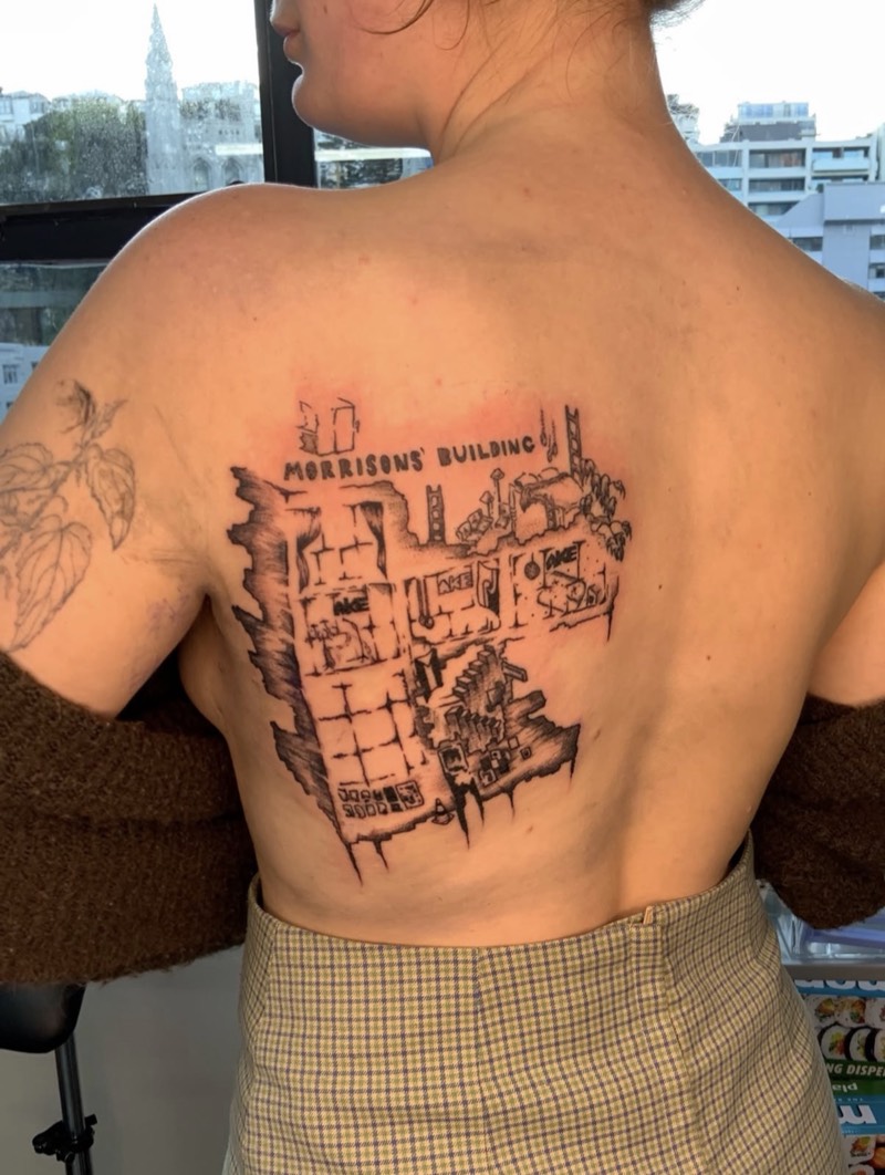 Completed tattoo on the back: a crumbling building interior and city skyline in fine black line, under hand lettering reading "Morrisons' Building"