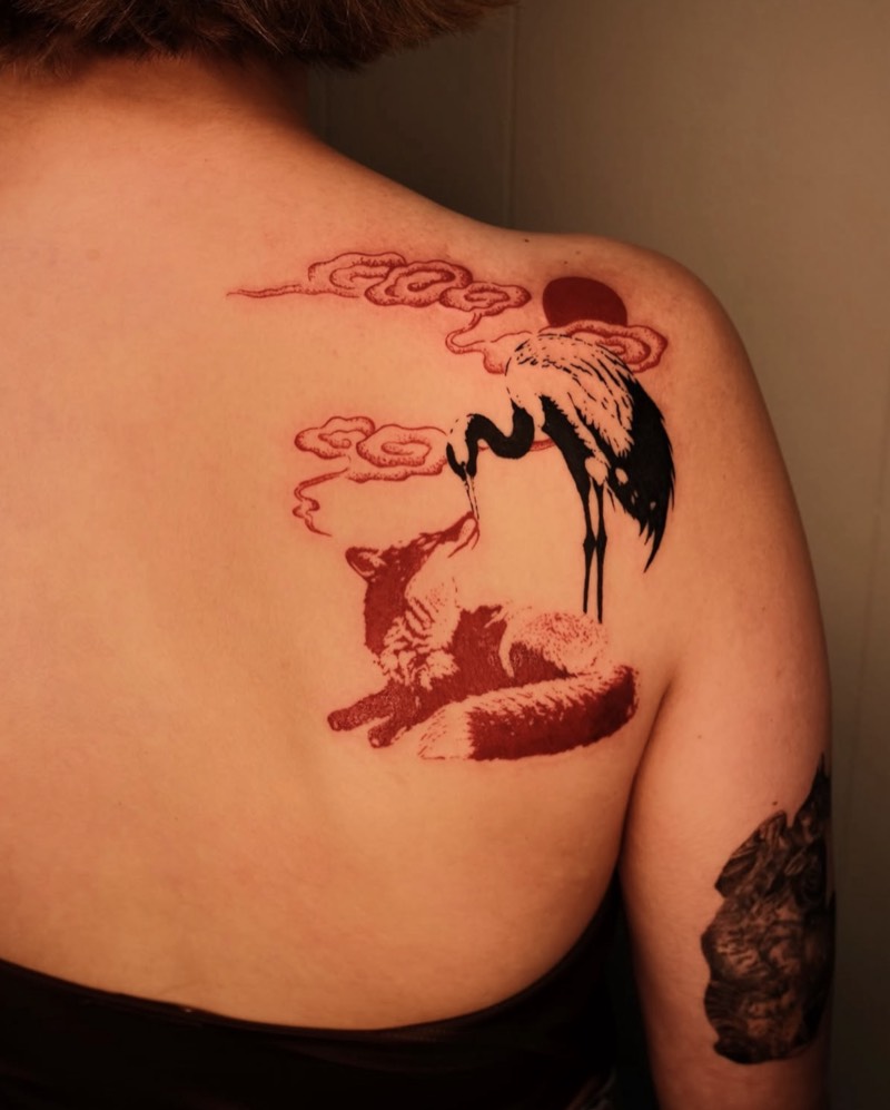 Completed tattoo on the shoulder blade: a red fox lying down and looking up at a black crane, under red clouds and a red sun
